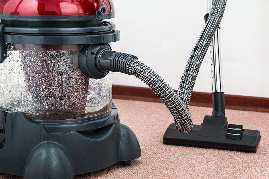 Close-up of a person operating a yellow portable carpet cleaning machine on a patterned rug in a well-lit room, with visible black ventilation grilles on the device. The individual's hand is pressing 