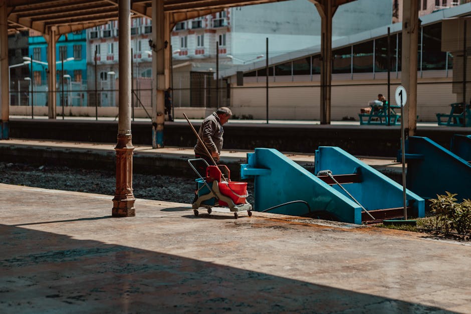 A man with gray hair wearing a gray jacket and jeans stands on a train station platform near a blue staircase leading down to the tracks, with a red stroller nearby. The platform is covered by a wooden canopy with supporting columns, and the ground is paved with concrete that appears clean and well-maintained. In the background, there are modern buildings with large windows, and a person sitting on a bench in the distance. The scene is illuminated by natural daylight, highlighting the tidy appearance of the area. This image emphasizes public space maintenance and cleanliness at a train station, aligning with the theme of surface cleaning and hygiene relevant to residential and commercial cleaning services by Carpet Cleaning Belsize Park.