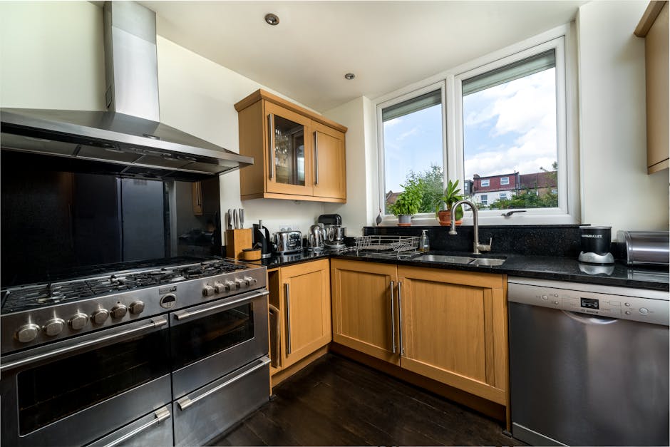 A modern kitchen with natural light streaming through two large front-facing windows, highlighting a black granite countertop and a stainless steel sink with a curved faucet. Wooden cabinets with a light oak finish are positioned beneath the countertop, alongside matching wall-mounted units, some with glass doors displaying dishware inside. On the countertop, there are small appliances including a toaster, kettle, and coffee maker, as well as potted green plants adding a touch of freshness. A stainless steel dishwasher is integrated into the cabinetry, and the black tiled backsplash contrasts with the light-colored walls. The dark hardwood floor provides a polished, clean look, indicating recent surface cleaning and maintenance. The scene reflects a well-maintained, hygienic space suitable for thorough domestic cleaning and deep cleaning routines, aligned with the end of tenancy cleaning checklist for Eton Avenue landlords, as covered by Carpet Cleaning Belsize Park.