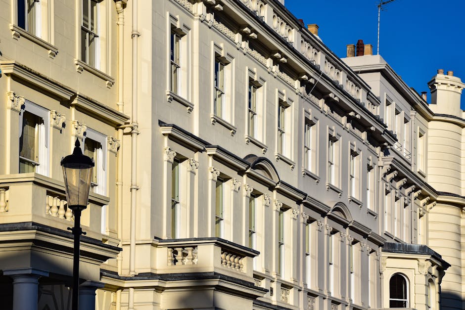 Photograph of a historic multi-storey building with cream-colored facades, adorned with decorative architectural details and large rectangular windows framed with white trim. The building features classic elements such as ornate cornices and balustrades, under bright daylight with a clear blue sky in the background. A black street lamp is visible in the foreground, standing beside the building. The scene highlights the exterior surface cleaning and preservation of the building's ornate features, emphasizing the importance of maintenance and visual hygiene in heritage properties, as promoted by Carpet Cleaning Belsize Park for residential and commercial properties, especially in preparing properties like Eton Avenue for end of tenancy cleaning.