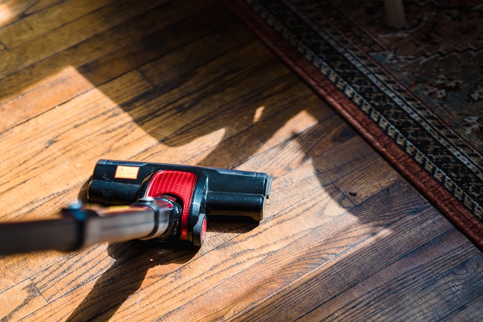 A handheld vacuum cleaner with a black body and red accents is positioned on a wooden floor near the edge of a patterned oriental rug. The scene is well-lit, highlighting the wood's natural grains and a clean, polished surface. The vacuum cleaner appears to be in use for surface cleaning or deep cleaning of the hardwood floor, demonstrating a typical domestic cleaning process. The environment suggests a tidy, well-maintained room, with the vacuum cleaner ready for sanitisation and routine cleaning tasks, as provided by Carpet Cleaning Belsize Park.
